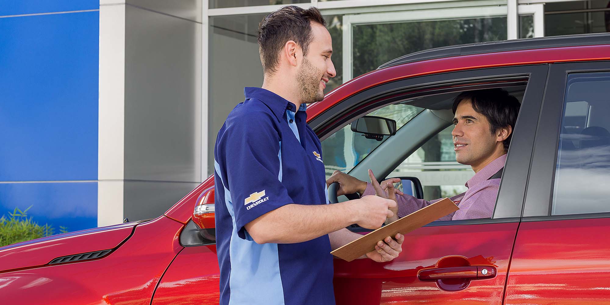 Asesor de servicio Chevrolet atendiendo a un cliente en su vehículo rojo